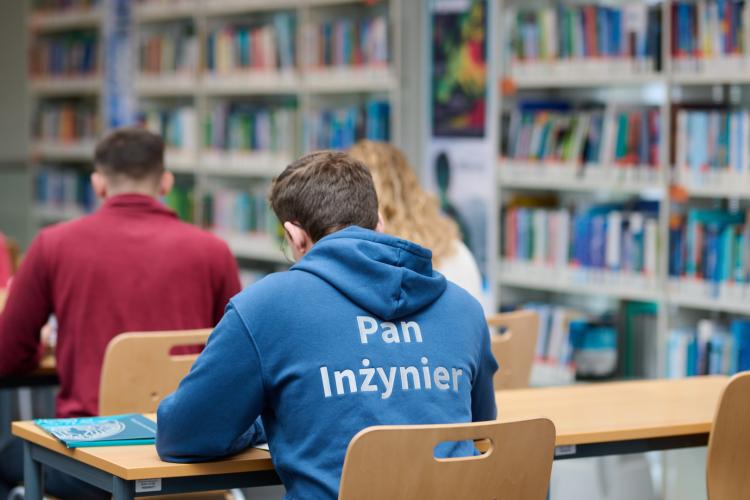 Na zdjęciu trzy osoby siedzące w bibliotece, widoczne tyłem 