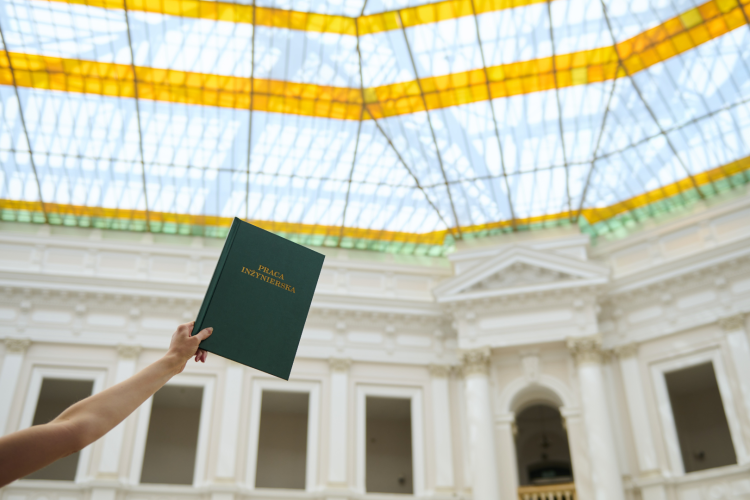 Zdjęcie przedstawia rękę trzymającą w górze zieloną okładkę pracy inżynierskiej na tle świetlika w Dużej Auli Gmachu Głównego Politechniki Warszawskiej.