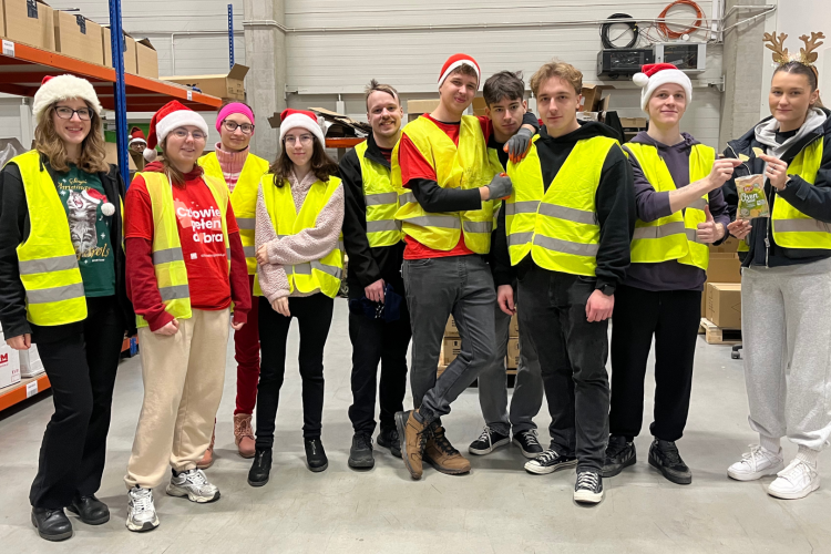 Zdjęcie przedstawia grupę 10 studentów, członków Studenckiego Koła Logistyki Stosowanej. Ubrani są w odblaskowe kamizelki, część z nich ma na sobie czapki świąteczne. Studenci stoją w magazynie między regałami z kartonami podczas wolontariatu realizowanego w ramach akcji Szlachetna Paczka.