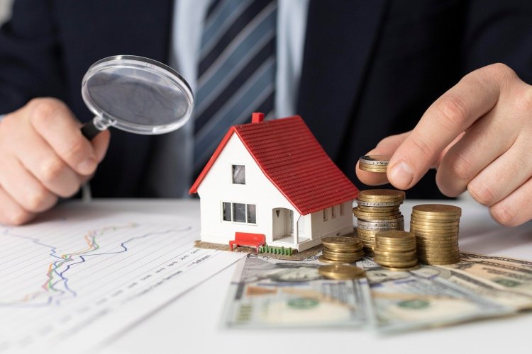 Na zdjęciu człowiek siedzący przy stole i trzymający lupę. Na stole leżą pieniądze i stoi figurka domu