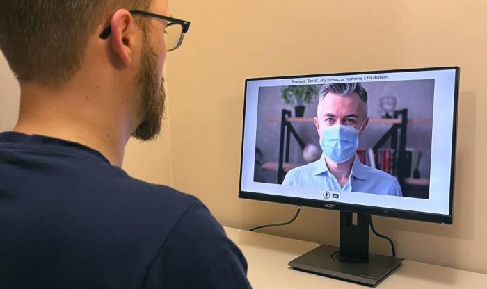 Zdjęcie mężczyzny przed ekranem monitora, na którym widoczna jest twarz innego mężczyzny w maseczce
