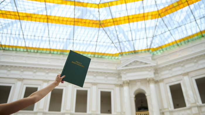 Zdjęcie przedstawia rękę trzymającą w górze zieloną okładkę pracy inżynierskiej na tle świetlika w Dużej Auli Gmachu Głównego Politechniki Warszawskiej.