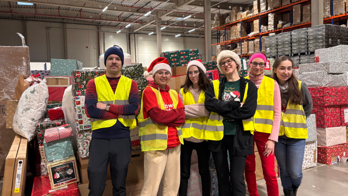 Zdjęcie przedstawia grupę 6 studentów, członków Studenckiego Koła Logistyki Stosowanej. Ubrani są w odblaskowe kamizelki, część z nich ma na sobie czapki świąteczne. Studenci stoją w magazynie między regałami z kartonami podczas wolontariatu realizowanego w ramach akcji Szlachetna Paczka.