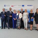 Na zdjęciu grupa stojących osób: przedstawiciele Politechniki Warszawskiej, Sopockiej Akademii Nauk Stosowanych, Zachodniopomorskiego Uniwersytetu Technologicznego w Szczecinie wraz z Ministrem Nauki Marcinem Kulaskiem i Dyrektorem NCBR prof. Jerzym Małachowskim 