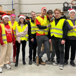 Zdjęcie przedstawia grupę 10 studentów, członków Studenckiego Koła Logistyki Stosowanej. Ubrani są w odblaskowe kamizelki, część z nich ma na sobie czapki świąteczne. Studenci stoją w magazynie między regałami z kartonami podczas wolontariatu realizowanego w ramach akcji Szlachetna Paczka.